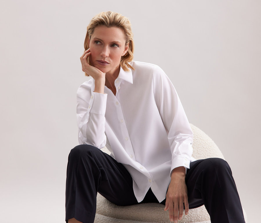 Žena sedí na židli, má na sobě bílou košili a černé kalhoty.