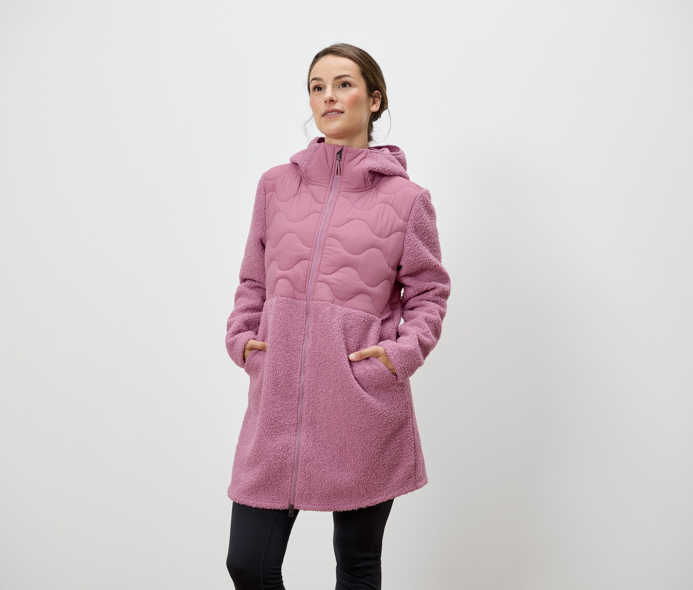 Žena stojí a má ruce v kapsách růžového kabátu s kapucí na zip.