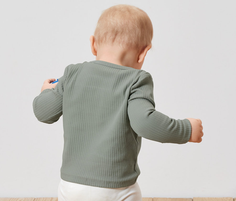 Batole zády ke kameře má na sobě světle zelené žebrované tričko s dlouhým rukávem a bílé kalhoty.