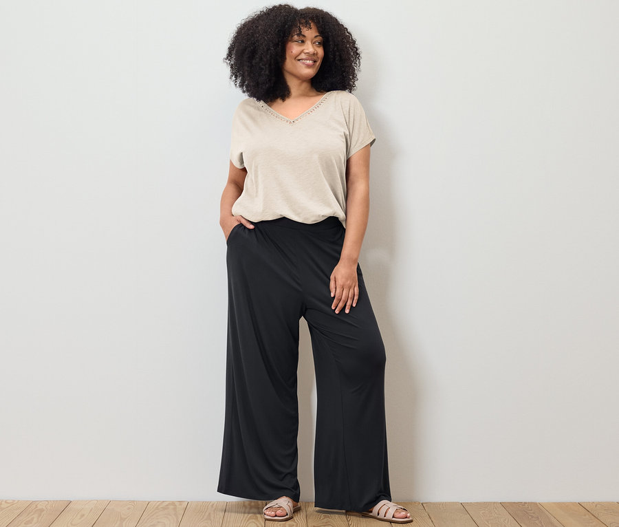 Žena pózuje v krémově bílém tričku z modalové směsi a černých kalhotách s širokými nohavicemi.