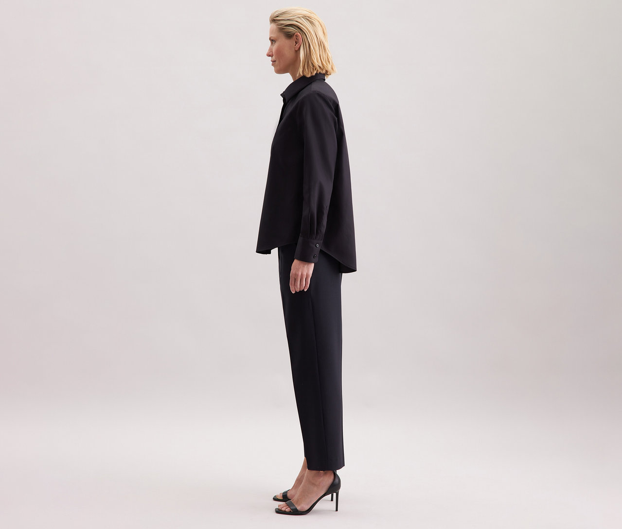 Žena má na sobě černou halenku Seidensticker, černé kalhoty a boty na vysokém podpatku.