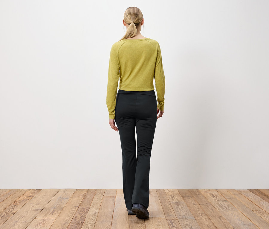 Žena s blond vlasy staženými do culíku stojí zády k fotoaparátu a má na sobě olivově zelený top s dlouhým rukávem a černé kalhoty.