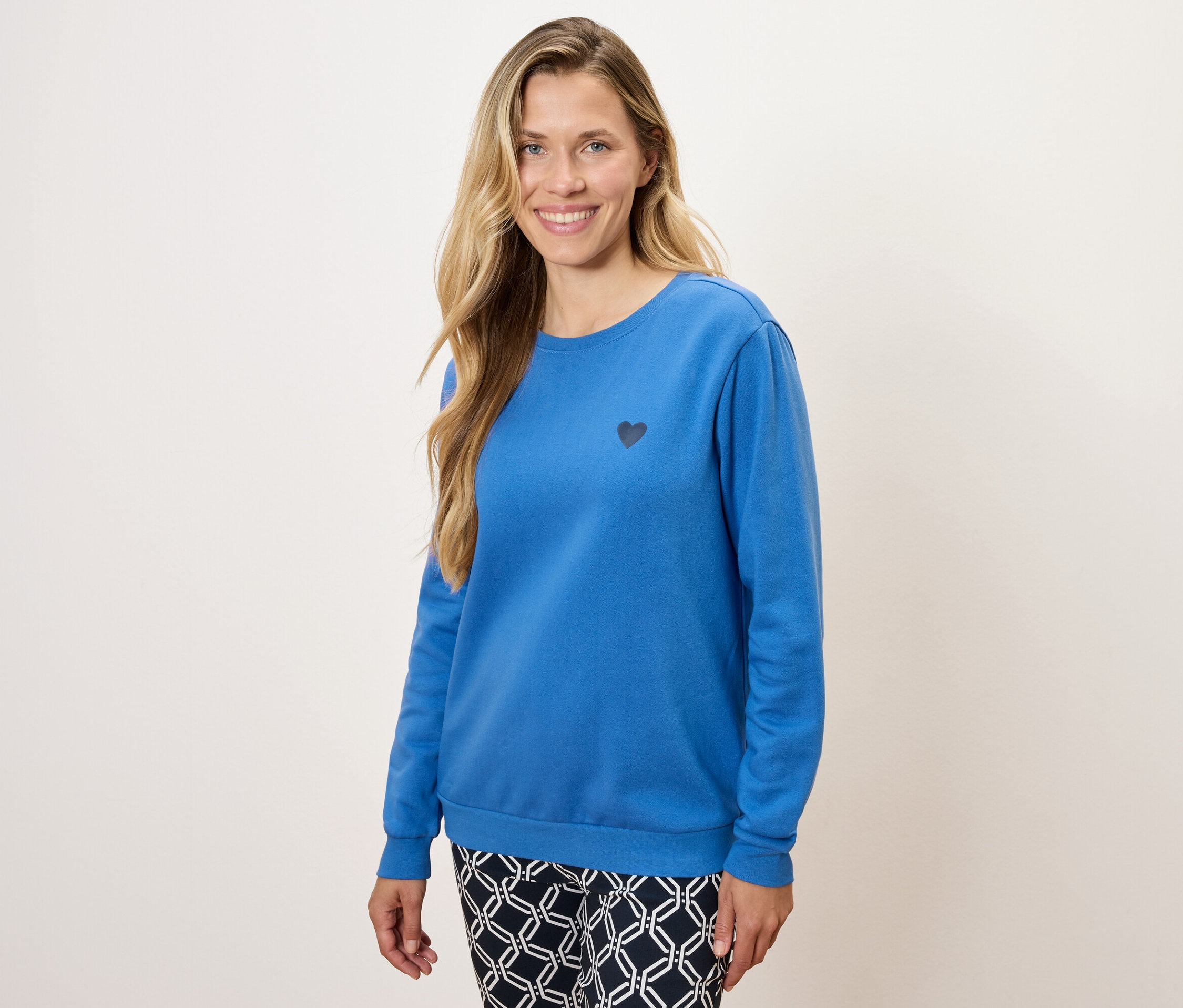 Žena má na sobě modrou mikinu s dlouhým rukávem a malým černým srdcem a vzorované kalhoty.