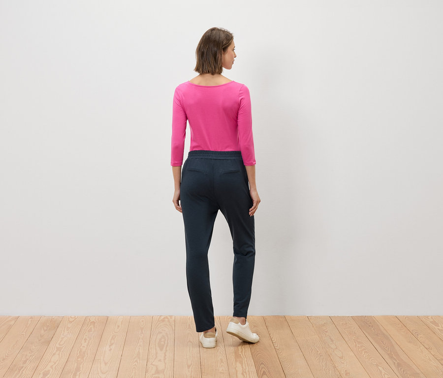 Žena stojí zády ke kameře, má na sobě růžovou halenku a modré kalhoty.