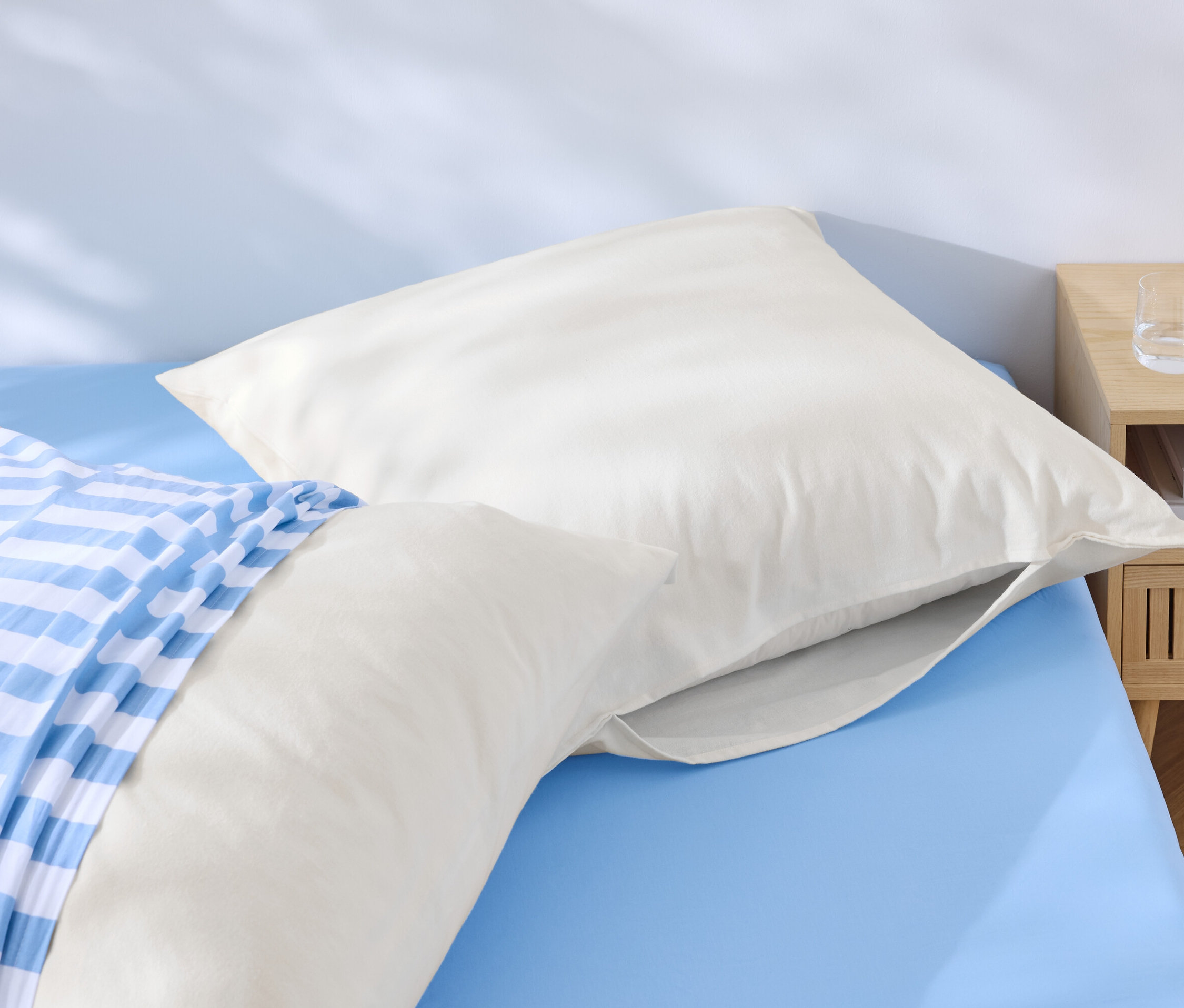 Dva bílé polštáře leží na modré posteli, jeden s modrobílým pruhovaným povlakem. Vedle postele stojí noční stolek s vodou a telefonem.