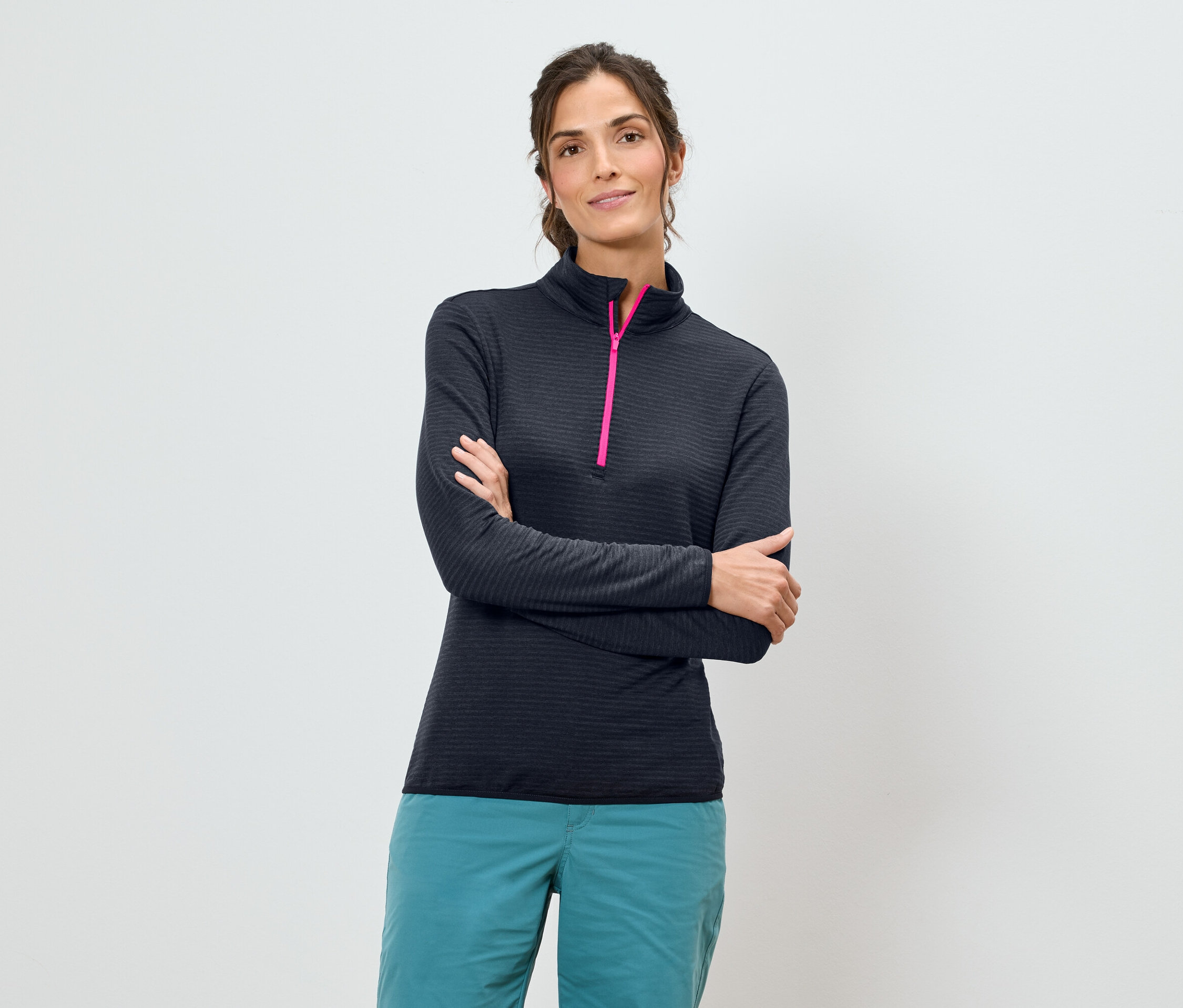 Žena se zkříženýma rukama má na sobě tmavě modrou košili s dlouhým rukávem s růžovým zipem a tyrkysové kalhoty na bílém pozadí.