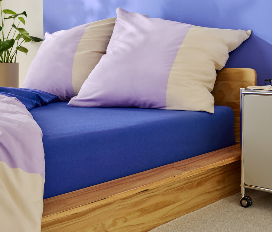 Postel s modrým prostěradlem a polštáři s fialovými a béžovými pruhy.