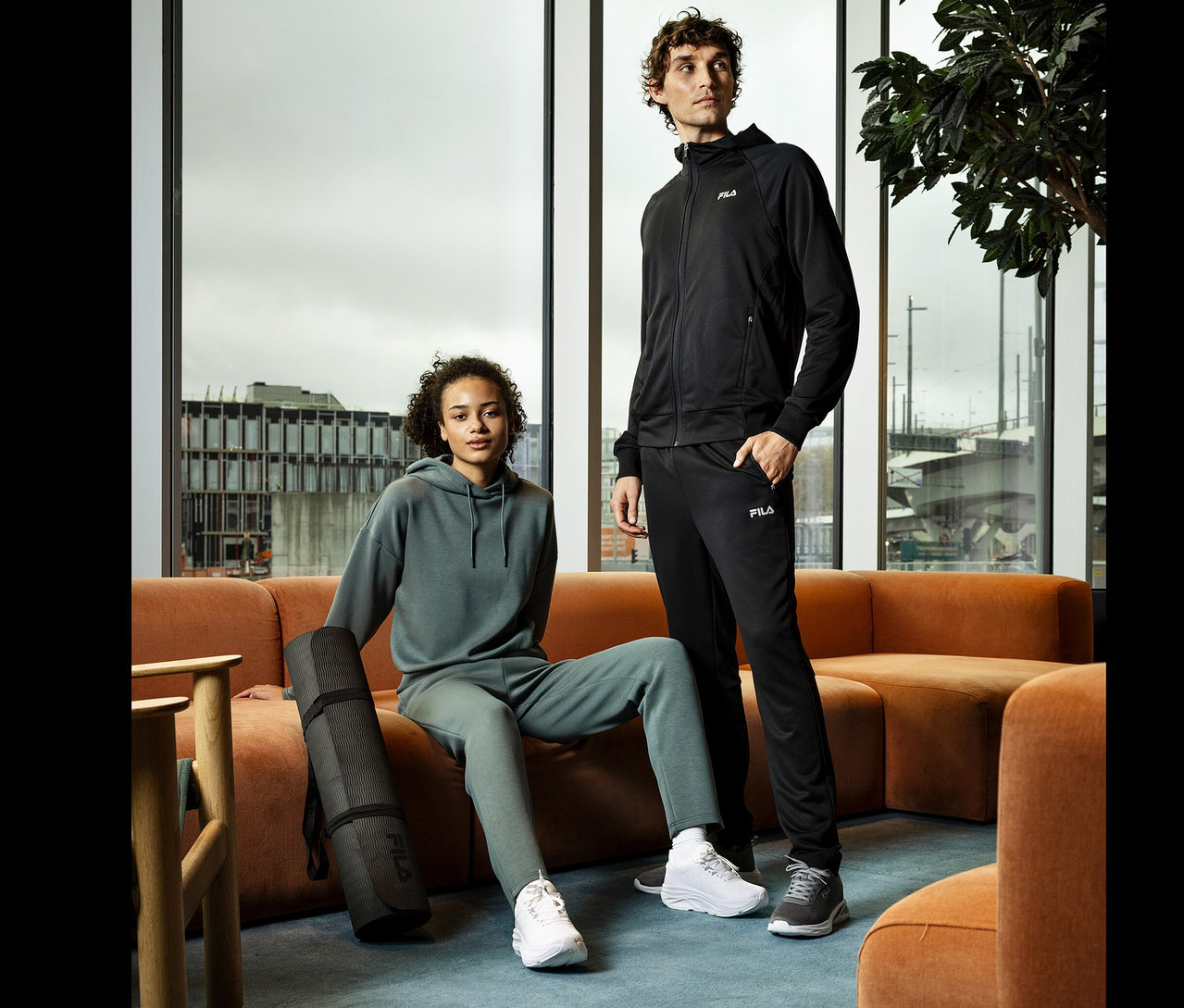 Žena sedí na oranžové pohovce s černou podložkou na jógu. Vedle ní stojí muž. Oba mají na sobě sportovní oblečení značky Fila.