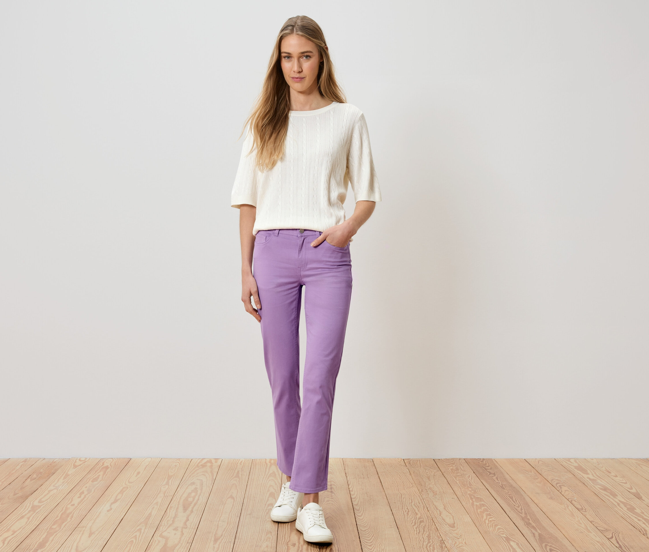 Žena stojí s rukou v kapse a má na sobě fialové kalhoty, bílý svetr a bílé boty.