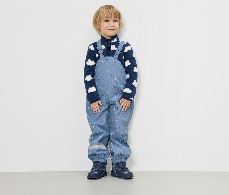 Malý chlapec stojí před bílou zdí a má na sobě modrou fleecovou bundu s obláčkovým vzorem a modré dešťové kalhoty s barevnými kapkami.