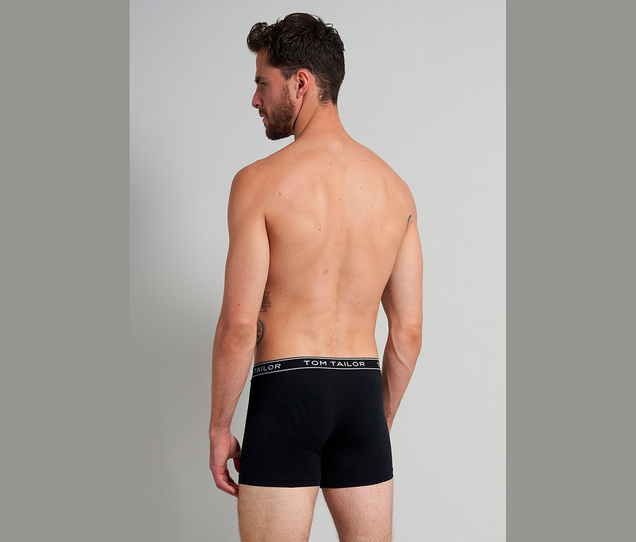 Muž zezadu, s tetováním, má na sobě černé boxerky Tom Tailor.