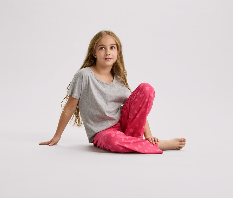 Dívka sedí na zemi v šedém tričku a růžových kalhotách s potiskem palem.