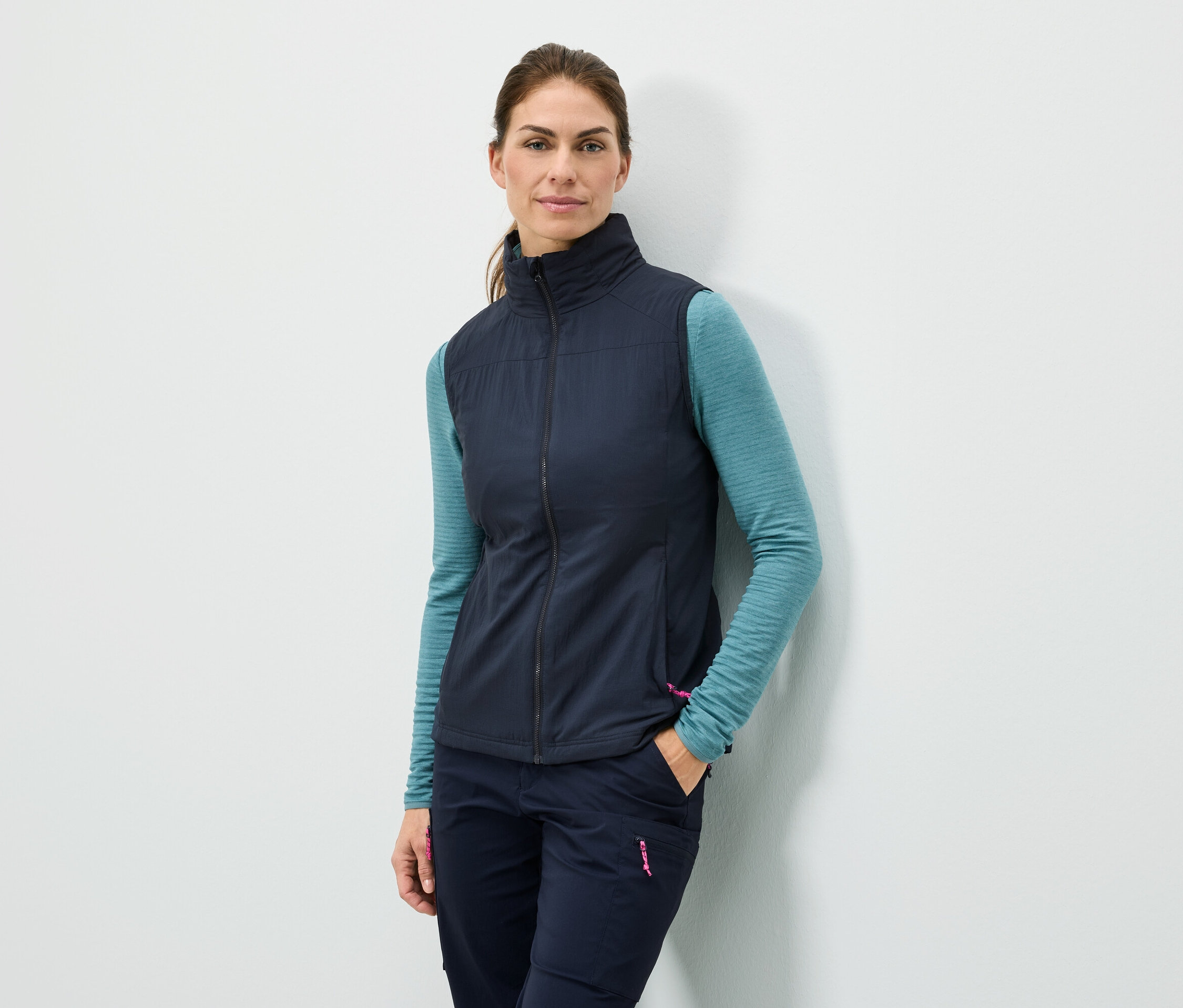 Žena stojí u zdi s rukou v kapse, má na sobě tmavě modrou vestu, tyrkysové tričko s dlouhým rukávem a tmavě modré kalhoty.