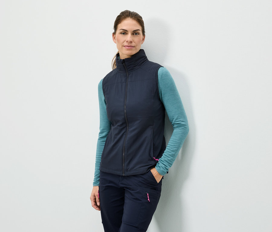 Žena stojí u zdi s rukou v kapse, má na sobě tmavě modrou vestu, tyrkysové tričko s dlouhým rukávem a tmavě modré kalhoty.