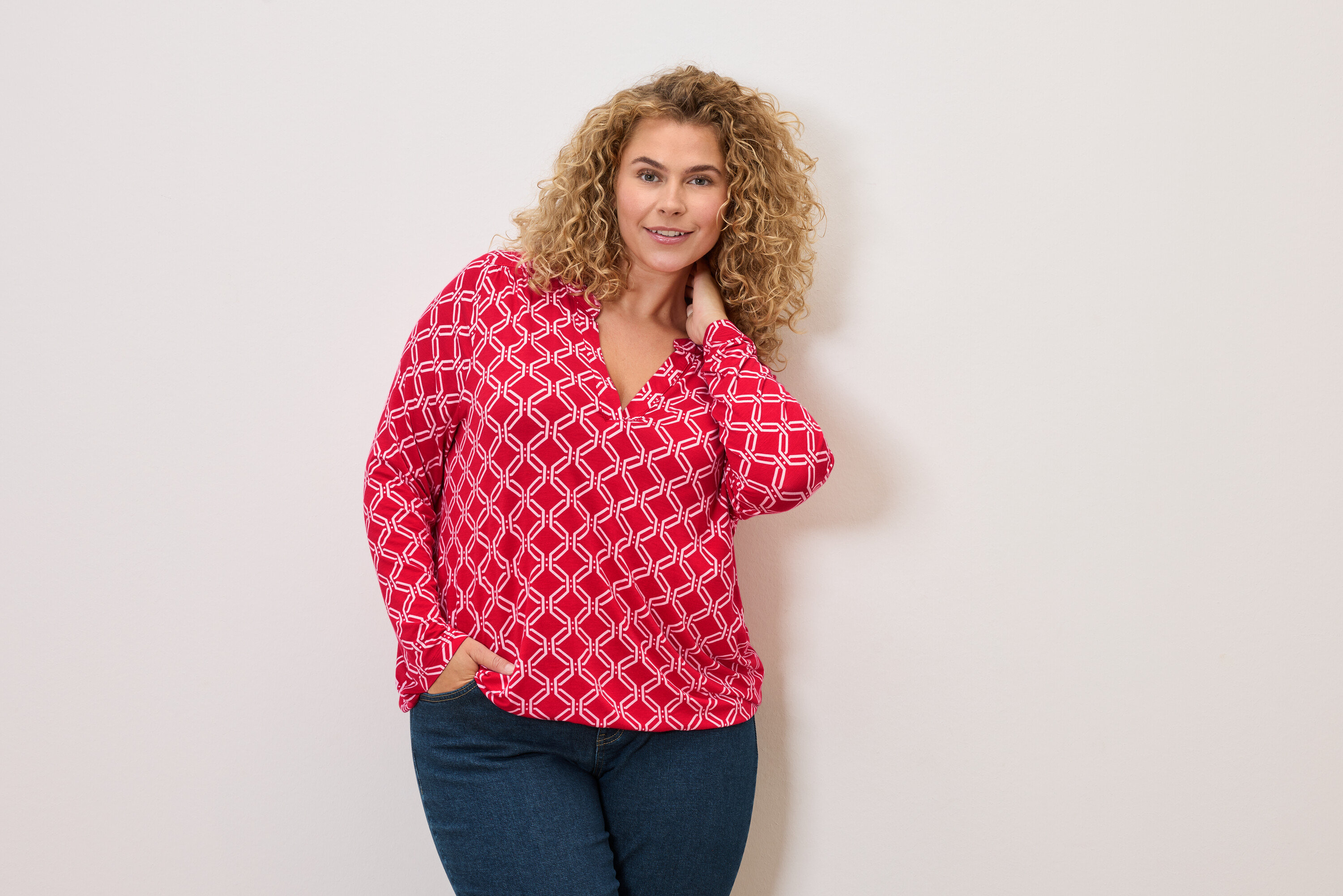Žena s kudrnatými vlasy v červenobílé halence a džínách pózuje s rukou v kapse.
