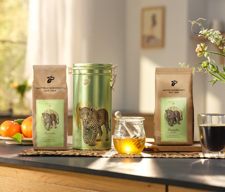 Na kuchyňské lince stojí dóza na kávu a dvě balení »Buramba Rwanda« včetně dózy na kávu vedle šálku kávy a medu.