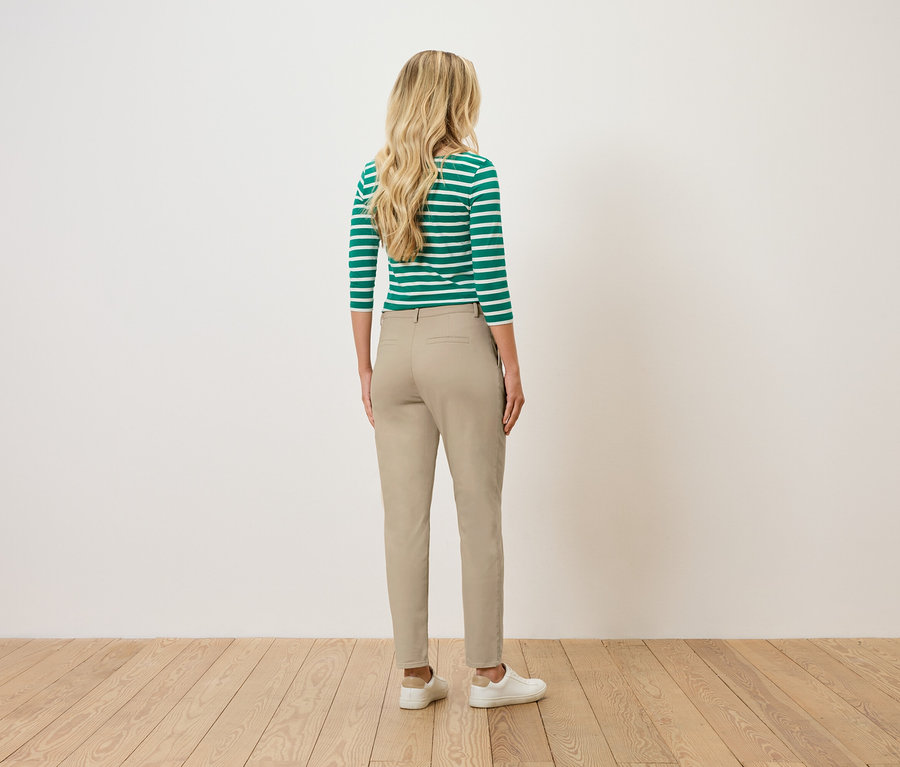 Žena s blond vlnitými vlasy stojí zády k divákovi a má na sobě zeleno-bílý pruhovaný top, béžové kalhoty a bílé tenisky.