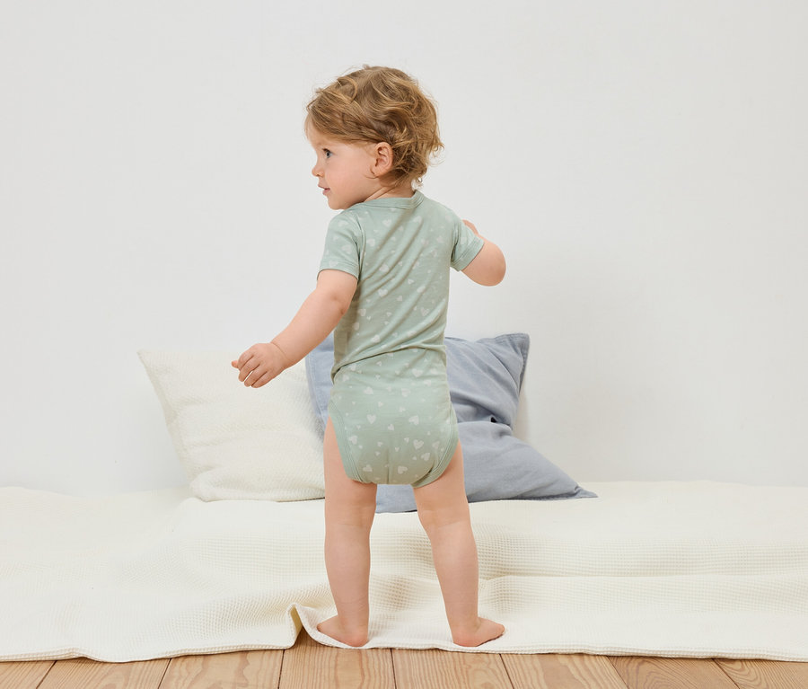 Malé dítě ve světle zeleném body s motivem srdce stojí na bílé hrací dece.