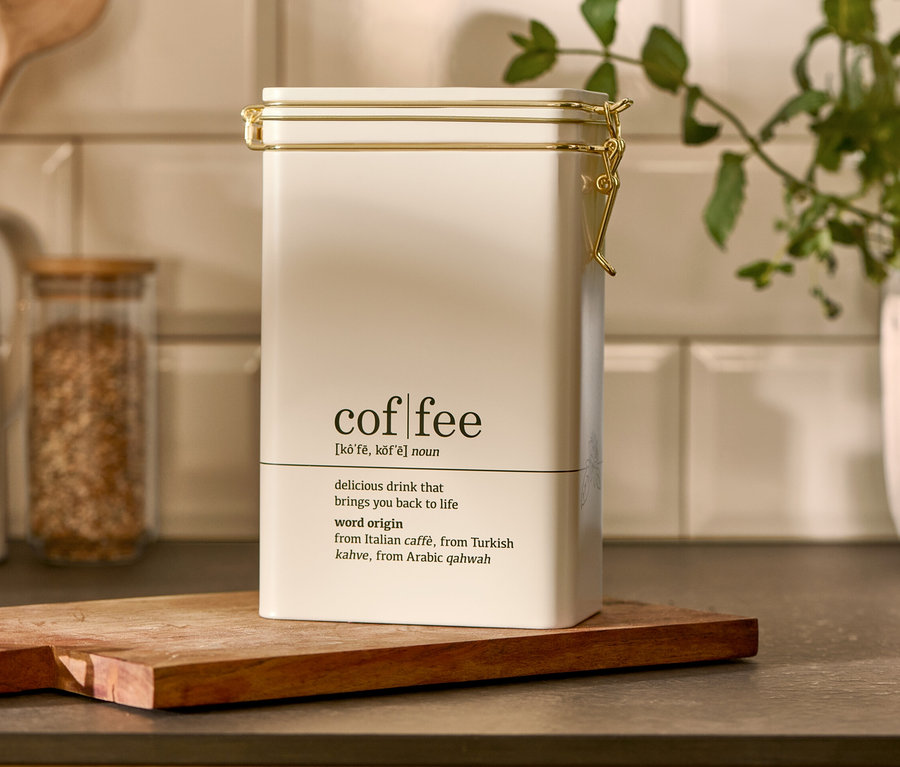 Bílá dóza na kávu "coffee dictionary" stojí na dřevěném prkénku.