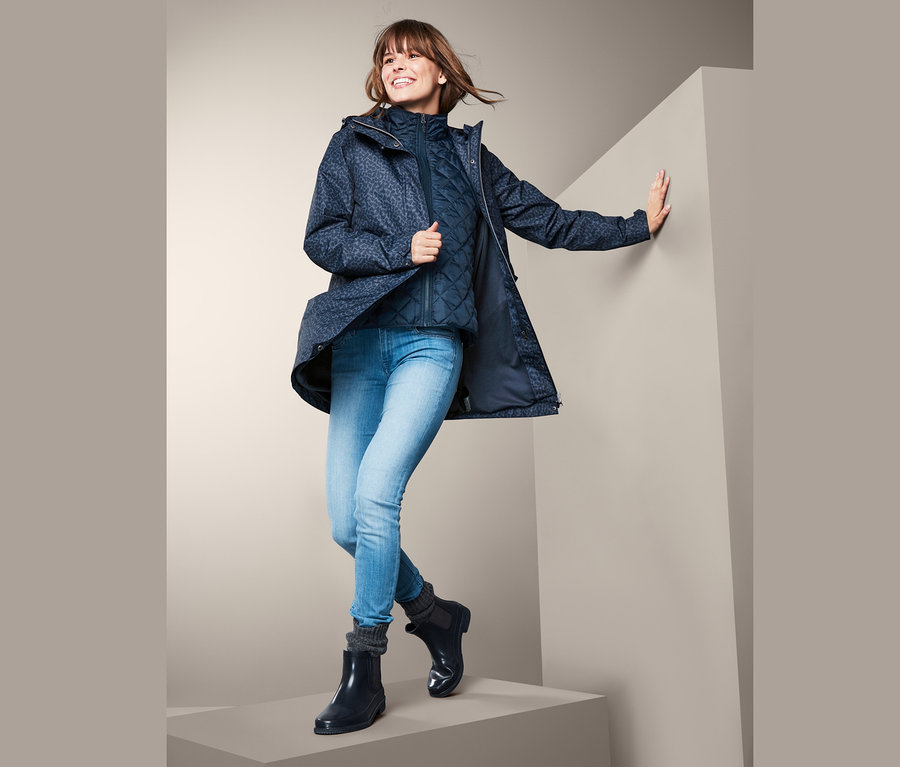 Usmívající se žena pózuje v bundě do deště 3 v 1, modré prošívané vestě, džínách a holínkách.