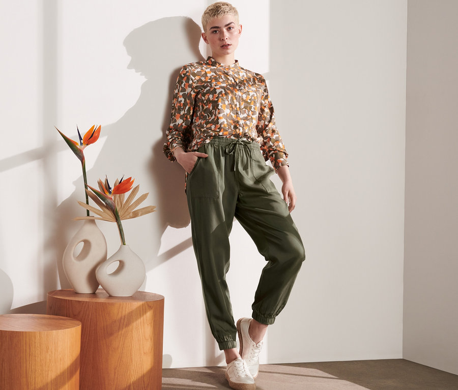 Žena pózuje opřená o zeď a má na sobě cargo kalhoty a vzorovanou košili.