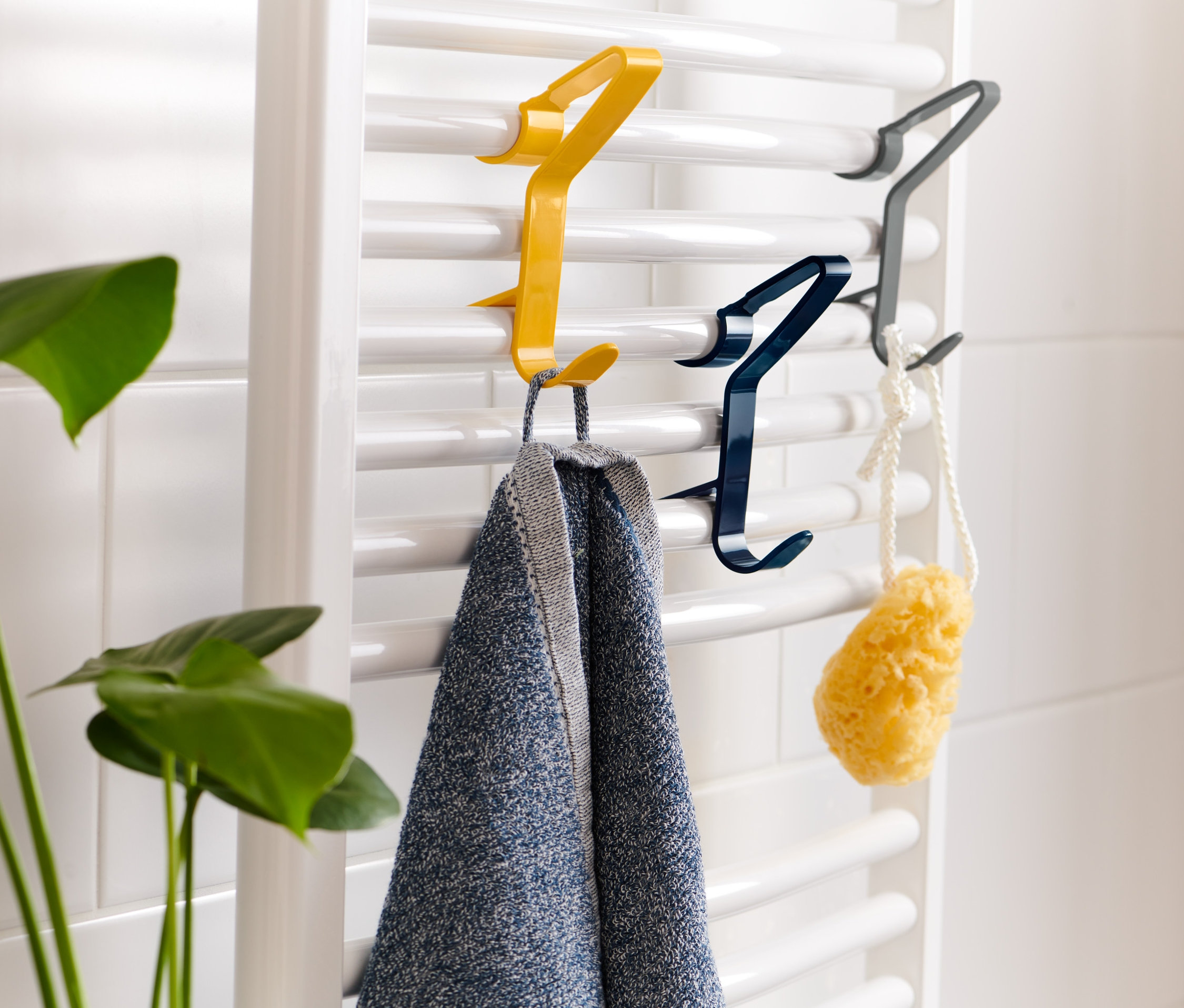 Na topení jsou zavěšeny 3 háčky na ručníky různých barev. Na žlutém háčku visí ručník, na modrém houba.