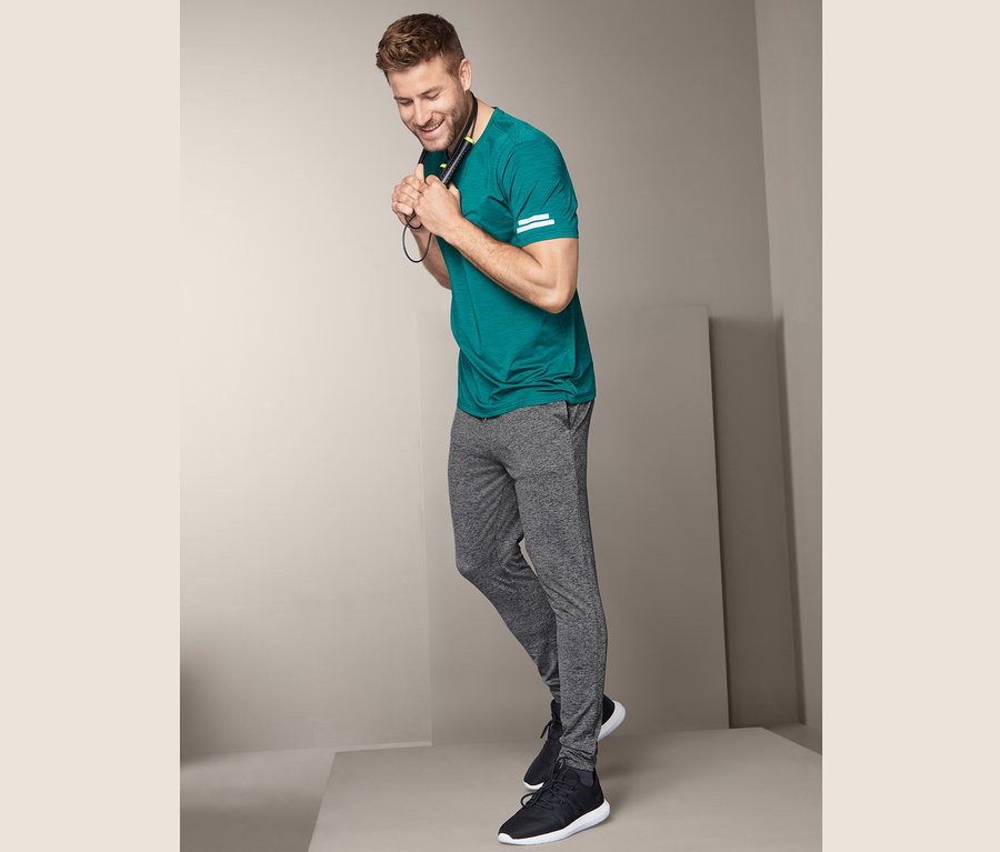 Muž v zeleném tričku drží sluchátka na krku a má na sobě sportovní kalhoty.