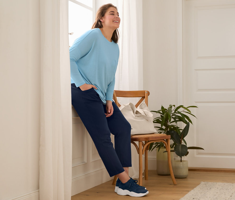 Žena opřená o okno má na sobě kožené boty, tmavě modré joggingové kalhoty, triko s netopýřími rukávy a plátěnou nákupní tašku.