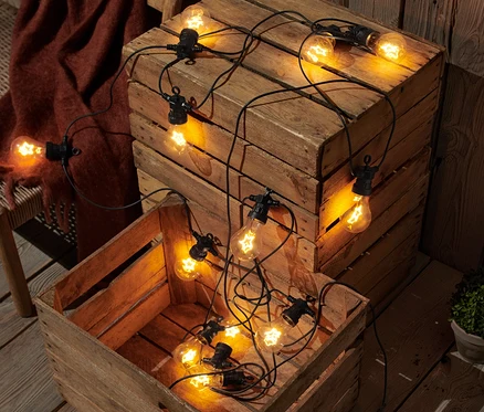 Dva dřevěné boxy s LED světelnými řetězy s vláknem, které leží na dřevěné terase vedle proutěné lavice s přehozenou hnědou dekou.
