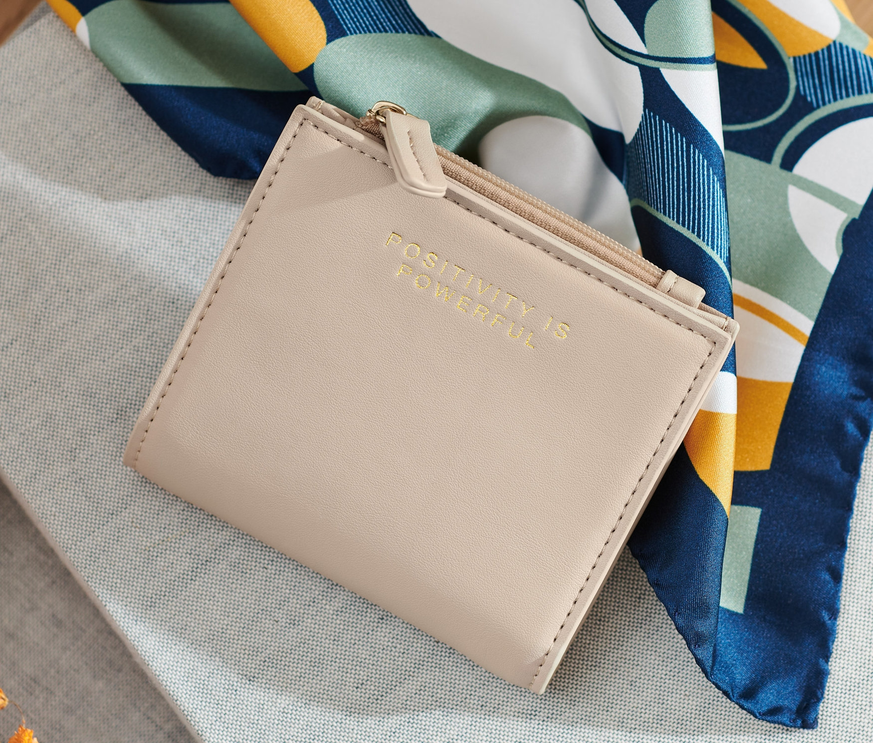 Krémová peněženka leží na šedé látce vedle květin a šátku.