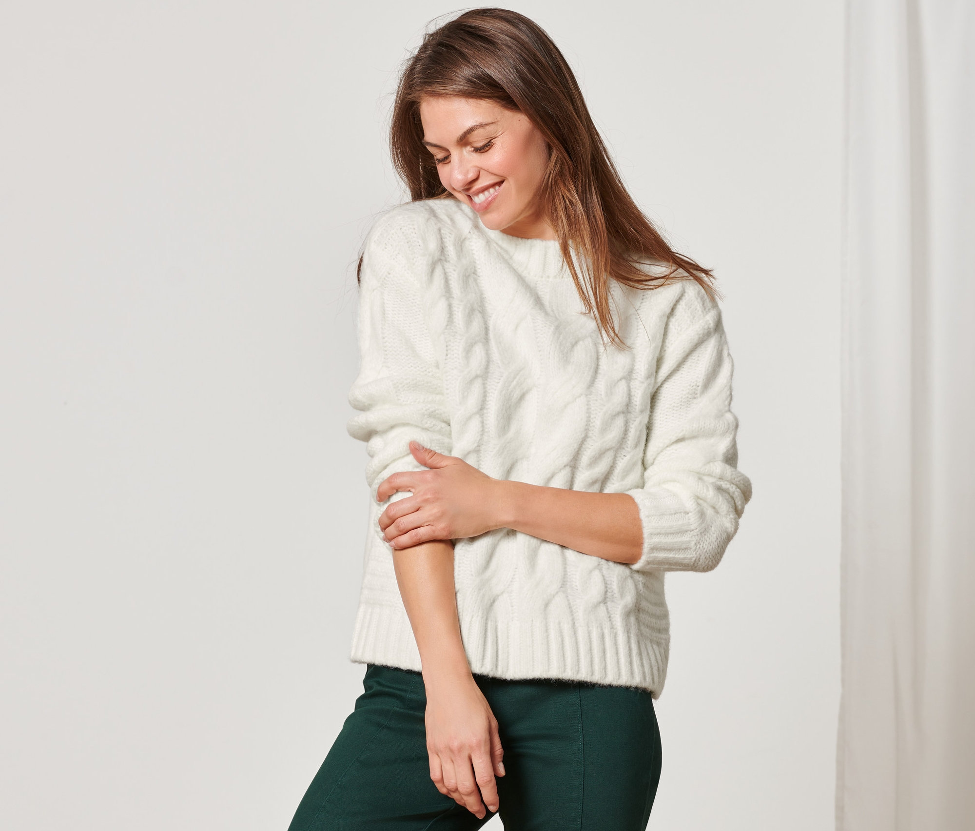 Žena v zelených kalhotách a pleteném svetru s copánkovým vzorem.