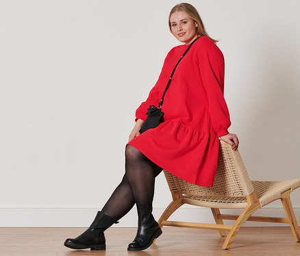 Žena sedí na proutěném křesle, má na sobě červené teplákové šaty, kožené boty, punčochové kalhoty (2 ks) a mini taštičky.