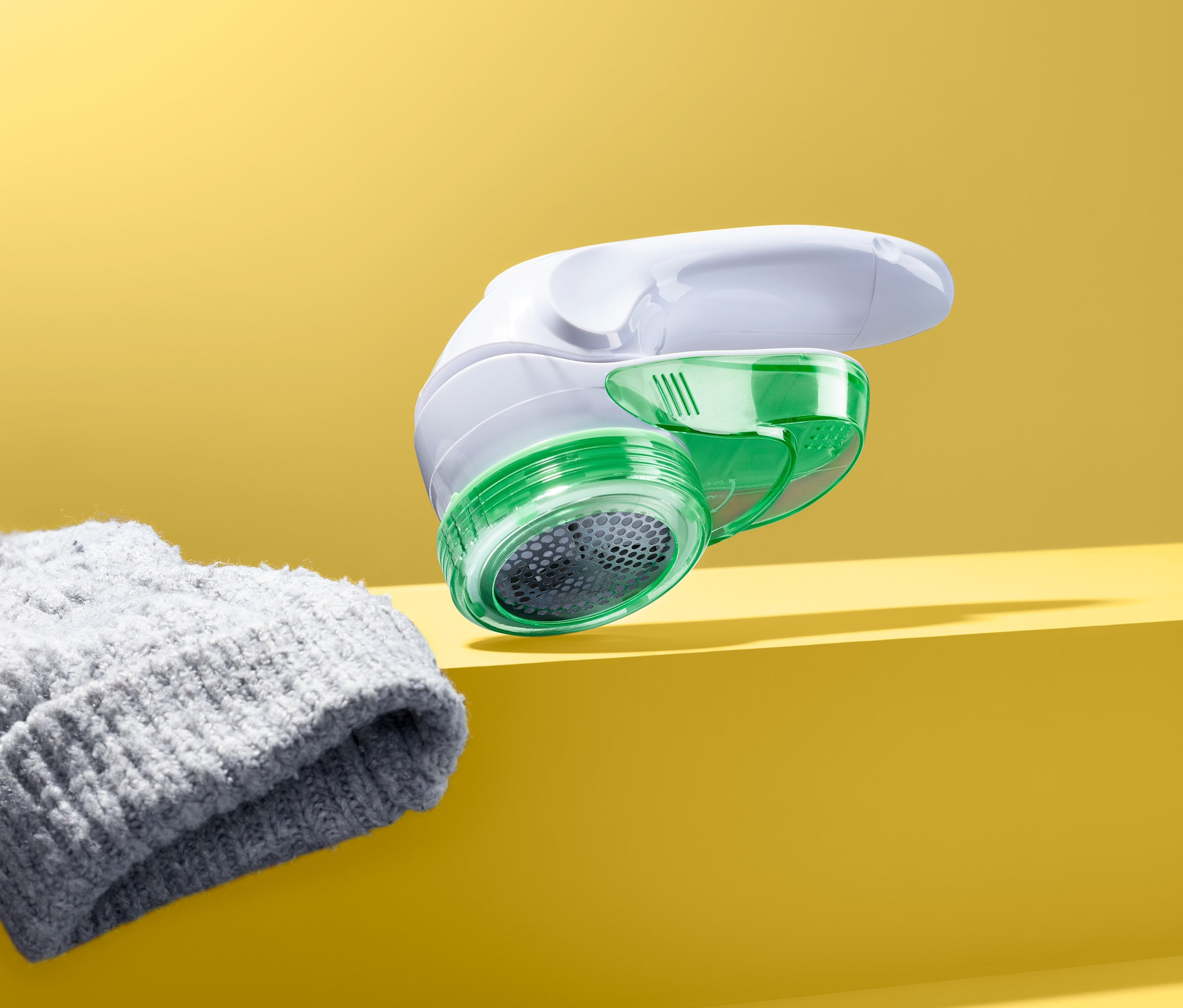 Odstraňovač žmolků s pohyblivou střihací hlavicí leží vedle šedé pletené čepice na žlutém pozadí.