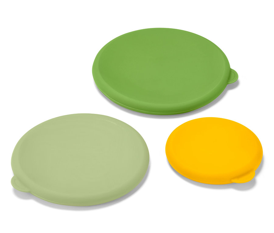 Tři silikonová víčka pro uchování čerstvosti, zelené, světle zelené a žluté barvy, leží na bílém povrchu.