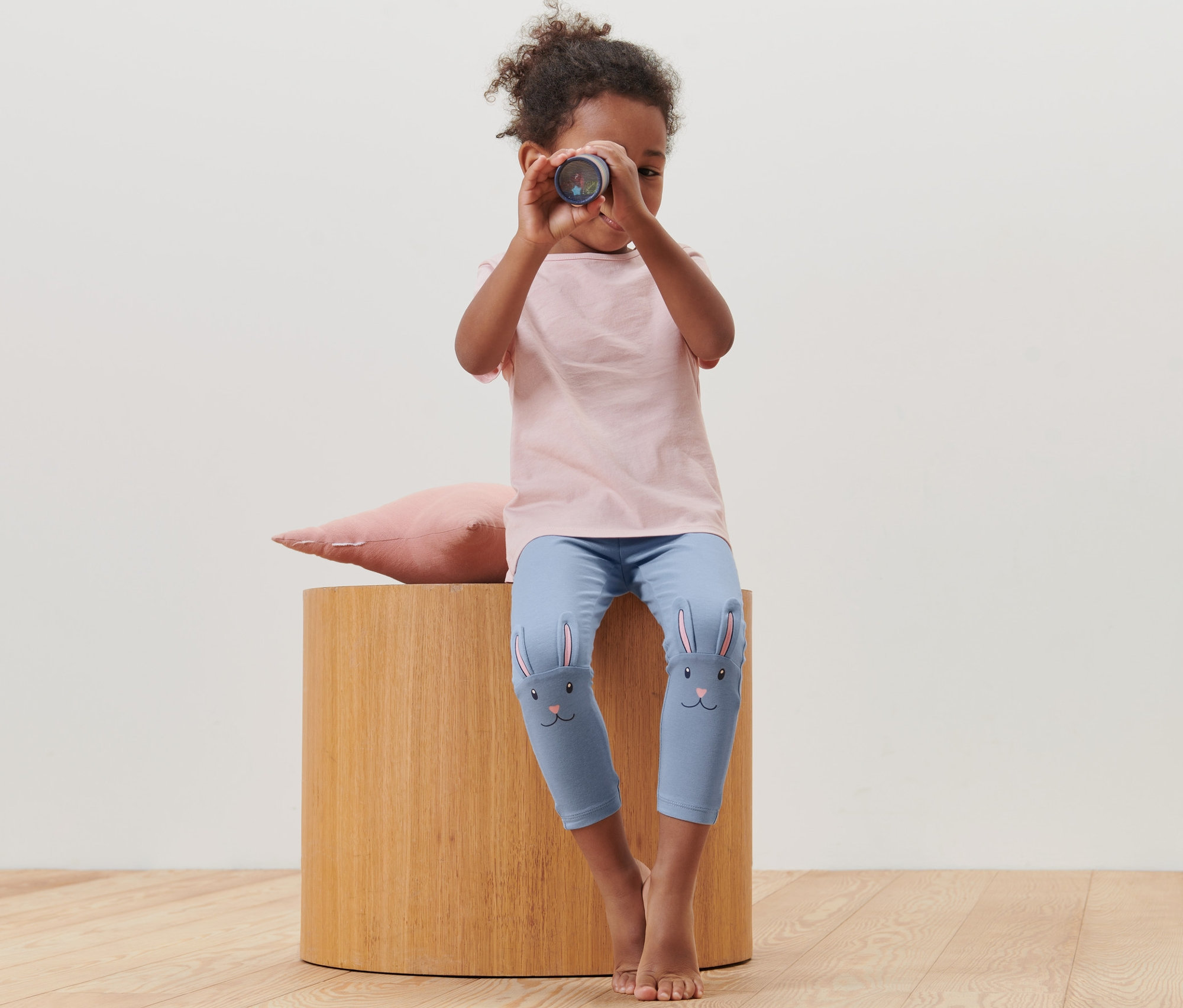 Dívka sedí na stoličce a dívá se dalekohledem. Má na sobě dětské legíny, 2 ks s motivem králíka.