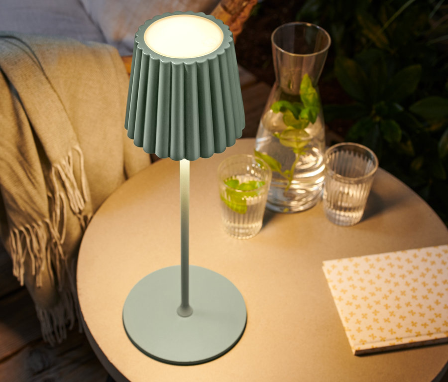 Na stole stojí zapnutá bateriová stolní lampa s LED v šalvějově zelené barvě.