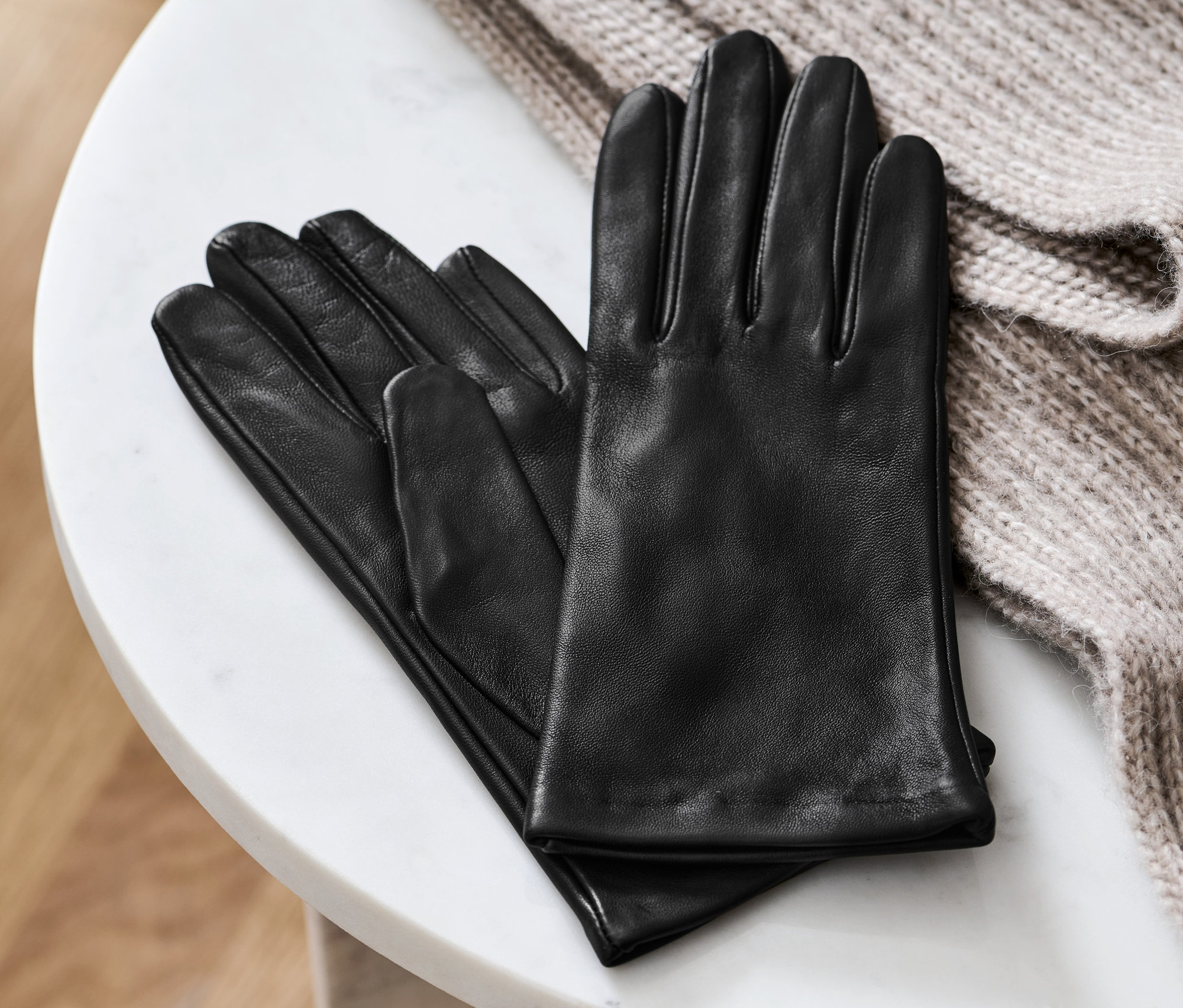 Černé kožené rukavice leží na bílém mramorovém stolku vedle béžového pleteného svetru.