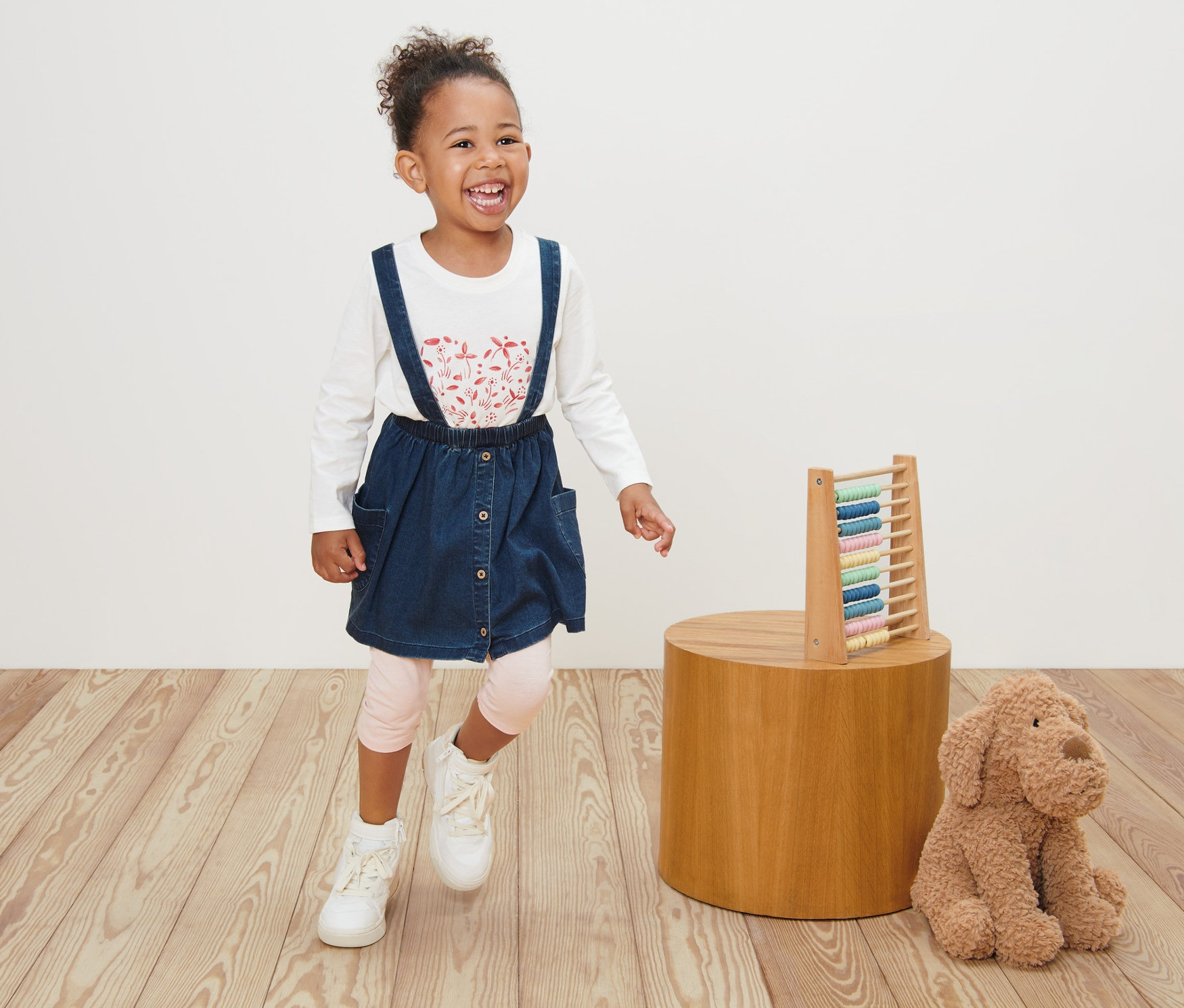 Usmívající se holčička v dětské džínové sukni a bílém tričku s dlouhým rukávem stojí na dřevěné podlaze vedle hračky.