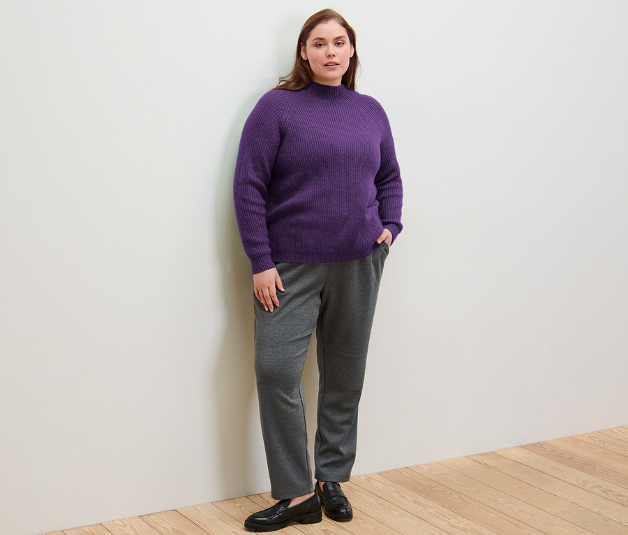 Žena stojí a má ruce v kapsách kalhot joggpant a na sobě má fialový svetr z hrubého úpletu se stojáčkem.