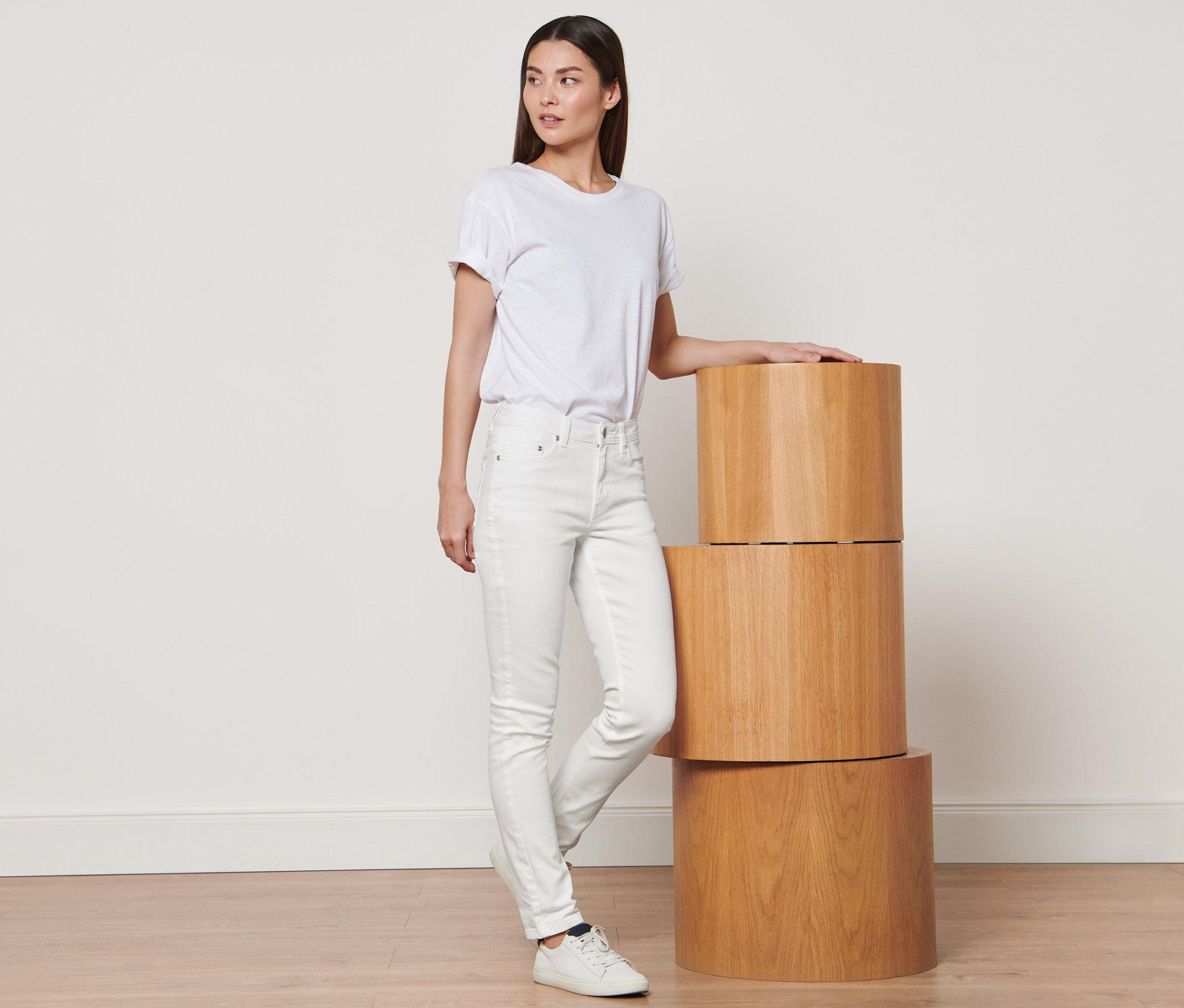 Žena stojí a opírá se o stoh tří dřevěných podstavců. Má na sobě bílé tričko a krémově bílé džíny »Fit Emma«.
