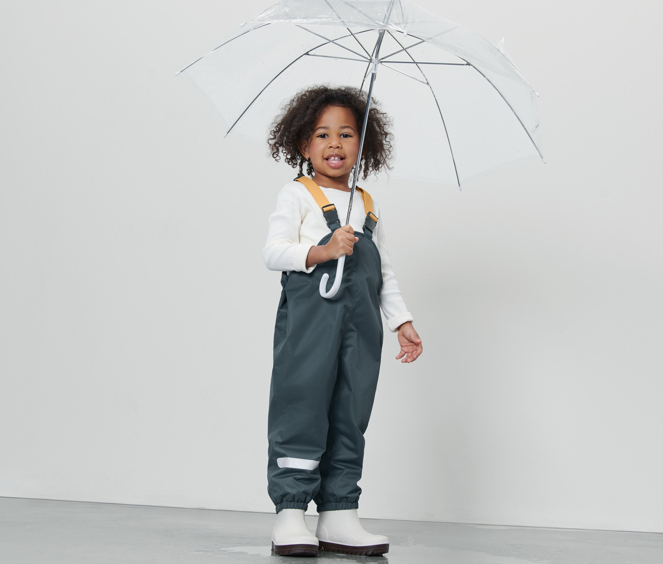 Dívka drží průhledný deštník, má na sobě dětské termo kalhoty do deště břidlicové modré, bílé boty a bílé tričko.