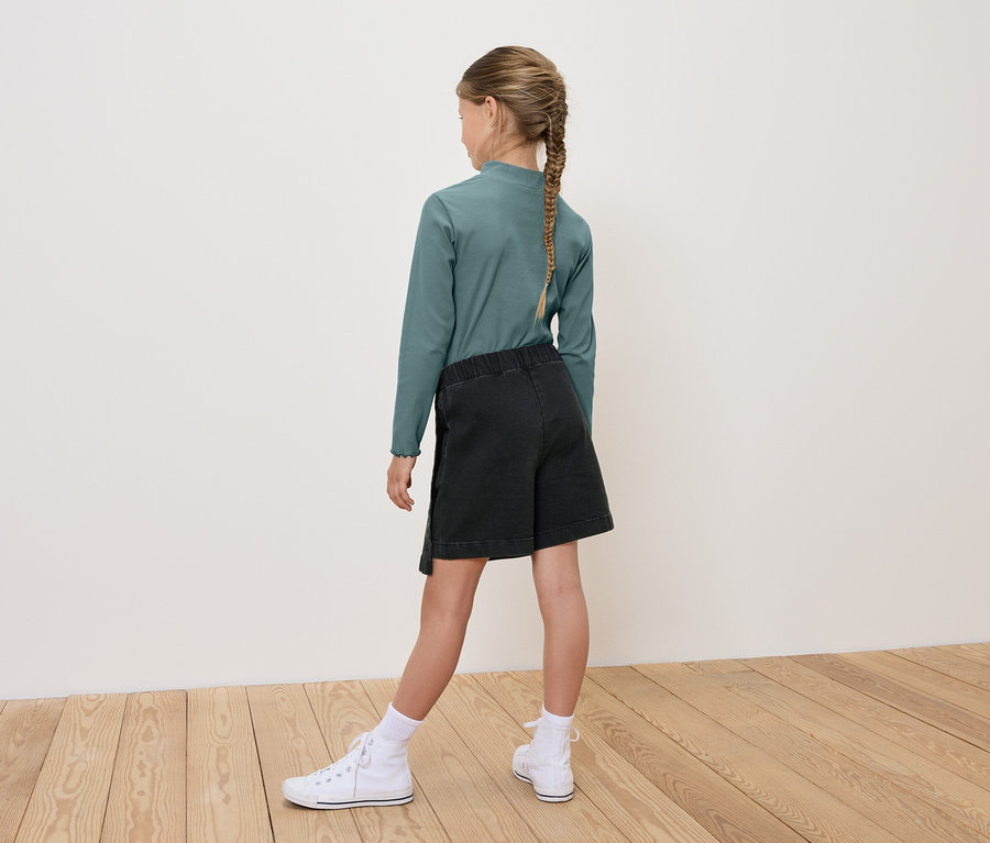 Dívka zády ke kameře má na sobě zelené tričko s dlouhým rukávem, černé šortky a bílé tenisky.