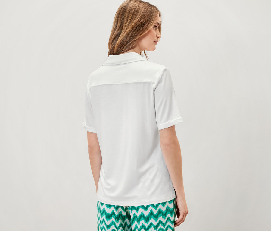 Žena stojí zády k fotoaparátu a má na sobě bílé halenkové tričko s knoflíkovou légou a zelené vzorované kalhoty.