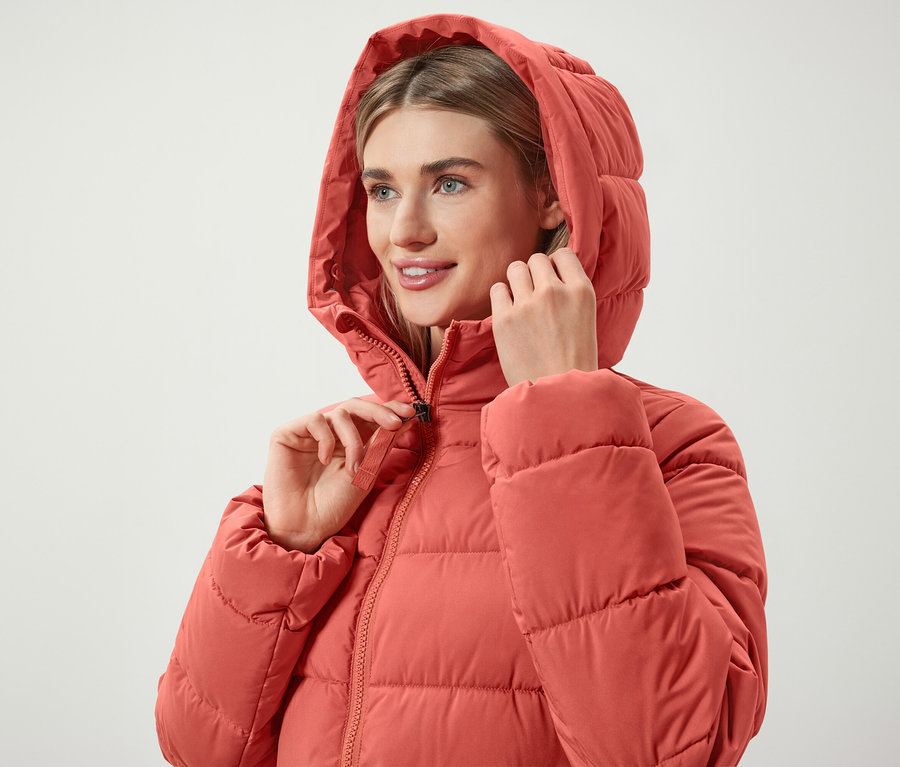 Žena si zapíná zip na korálově červené péřové bundě s kapucí.
