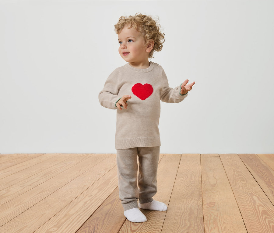 Batole s kudrnatými vlasy stojí na dřevěné podlaze a má na sobě béžový svetřík s červeným srdcem a béžové kalhoty.