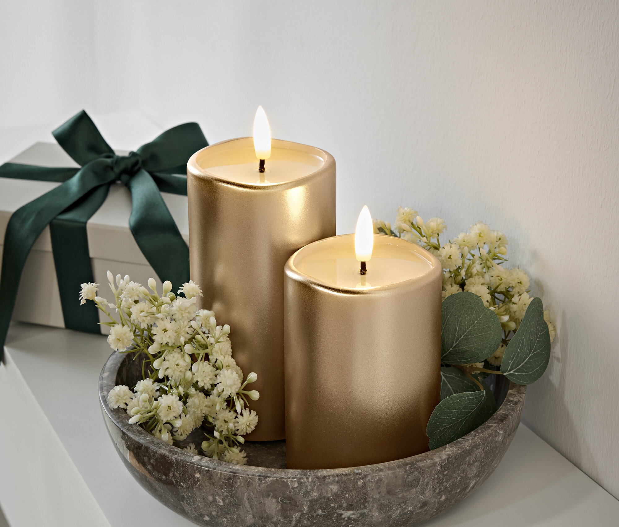 Dvě zlaté svíčky s plameny na šedé kamenné misce, bílé květy a zelené listy. Dárková krabička s tmavě zelenou stuhou v pozadí.