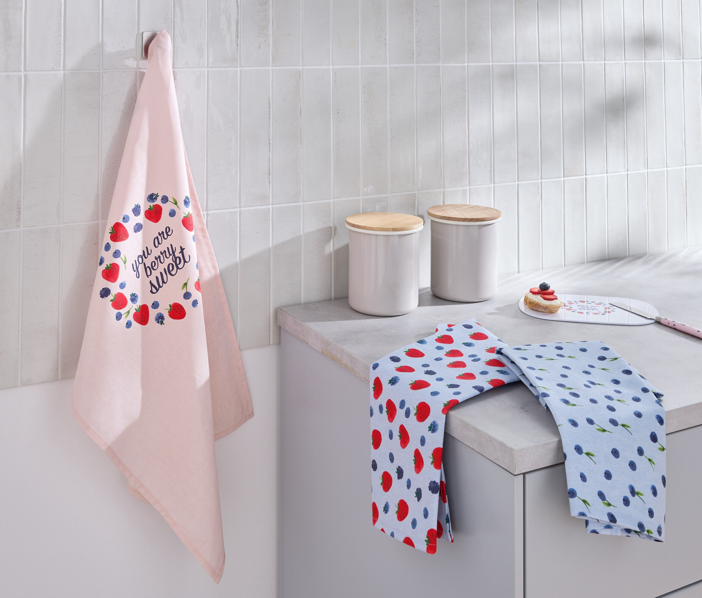 Na zdi visí světle růžová utěrka s potiskem jahod a borůvek. Na lince leží dvě složené utěrky a dva bílé kanystry s dřevěnými víčky.