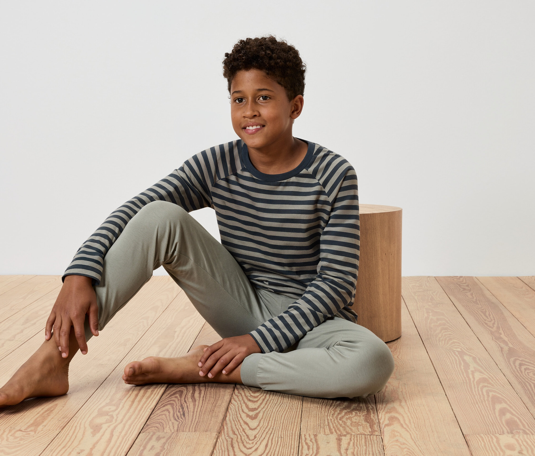 Mladý chlapec sedí na dřevěné podlaze, opírá se o malý dřevěný stolek, má na sobě pruhované tričko a kalhoty.