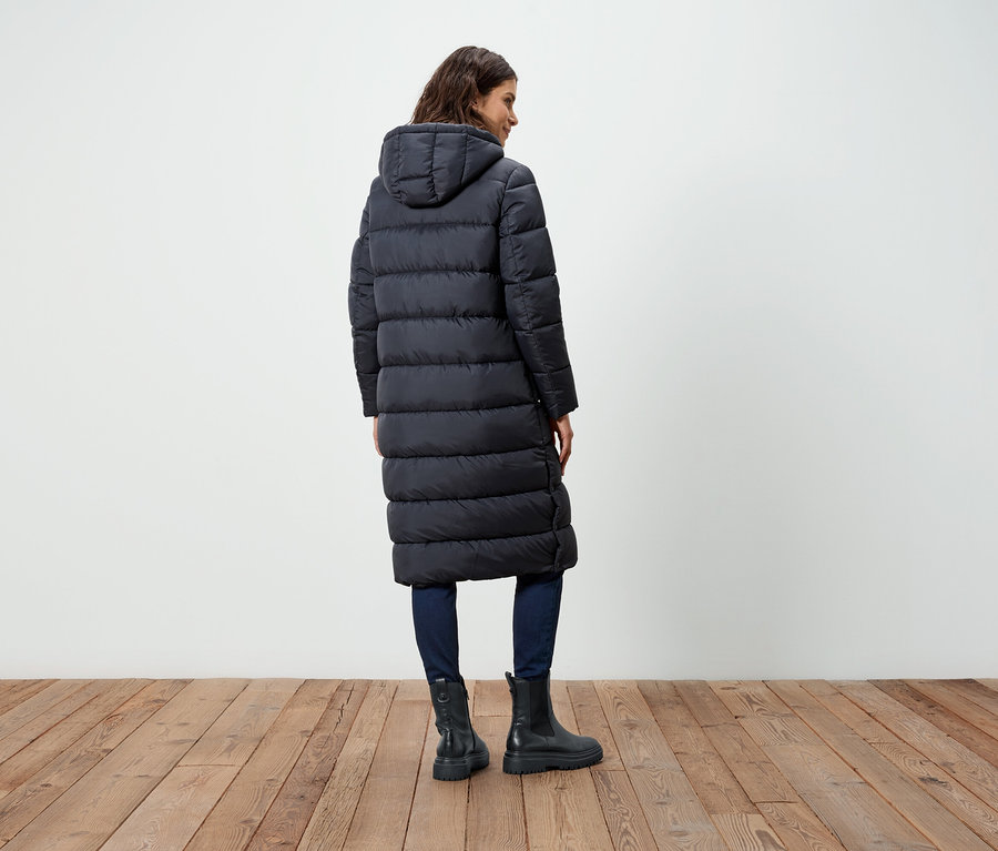 Žena zády k fotoaparátu má na sobě dlouhou černou péřovou bundu, džíny a boty.