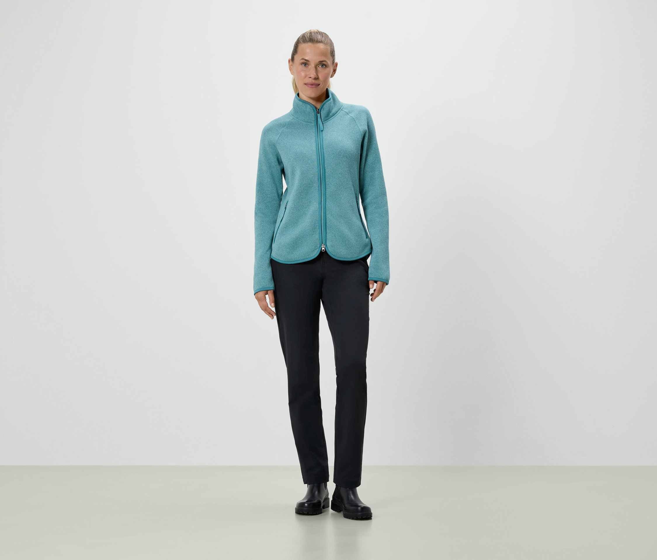 Žena stojí a má na sobě tyrkysovou fleecovou bundu a černé kalhoty.